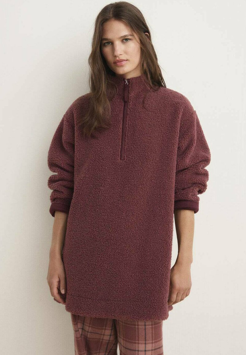 Pullover in pile bordeaux con colletto alto e zip a metà, caratterizzato da maniche oversize e una superficie testurizzata. Abbinato a pantaloni a quadri.