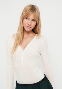 Ανοιχτό μπεζ μπλουζάκι μακρυμάνικο με V-λαιμόκοψη, διακοσμημένο με κουμπιά και μαλακό, υφασμάτινο ύφασμα. Συνδυάζεται με σκουρόχρωμη καρό φούστα.