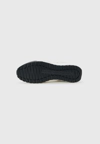 Zool van een sneaker met diepe groeven en Calvin Klein Jeans-logo, weergegeven tegen een eenvoudige witte achtergrond.