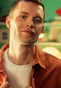 Ritratto di una persona che indossa una giacca arancione sopra una camicia bianca, con capelli corti e uno sfondo neutro nelle tonalità di verde.