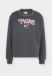 Mörkgrå Nike-tröja med rosa logotyp, swoosh och texten "Sedan 1972", med rosa kant runt kragen.