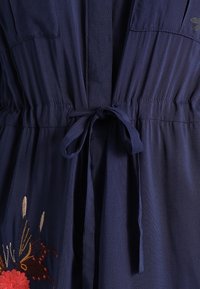 Vestido azul marino con un cinturón atado en la cintura y bordados florales en el lado inferior izquierdo.