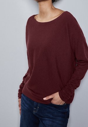 Vrouw draagt een donkerroze top met lange mouwen en gerimpelde mouwen, gecombineerd met een blauwe spijkerbroek, staand met één hand in de zak tegen een grijze achtergrond.