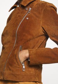 Braune Wildlederjacke mit einem Reißverschluss vorne, Druckknopfkragen und Seitentaschen. Verfügt über eine taillierte Passform und eine strukturierte Oberfläche.