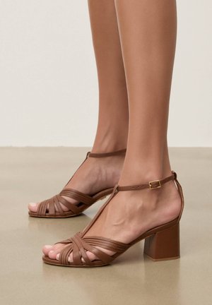 Fødder iført brune læder-sandaler med åben tå, ankelstropper og lave blokhæle på en glat beige gulv.