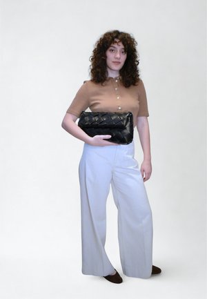 Bolso de mano negro tejido sostenido en la mano derecha, combinado con una blusa beige y pantalones anchos blancos. Cabello rizado y zapatos planos marrones. Fondo sencillo.