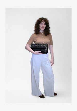 Μαύρο υφαντό τσαντάκι χειρός κρατημένο στο δεξί χέρι, φορεμένο με μπεζ τοπ και λευκό παντελόνι με φαρδιά πόδια. Σγουρά μαλλιά και καφέ επίπεδα παπούτσια. Απλό φόντο.