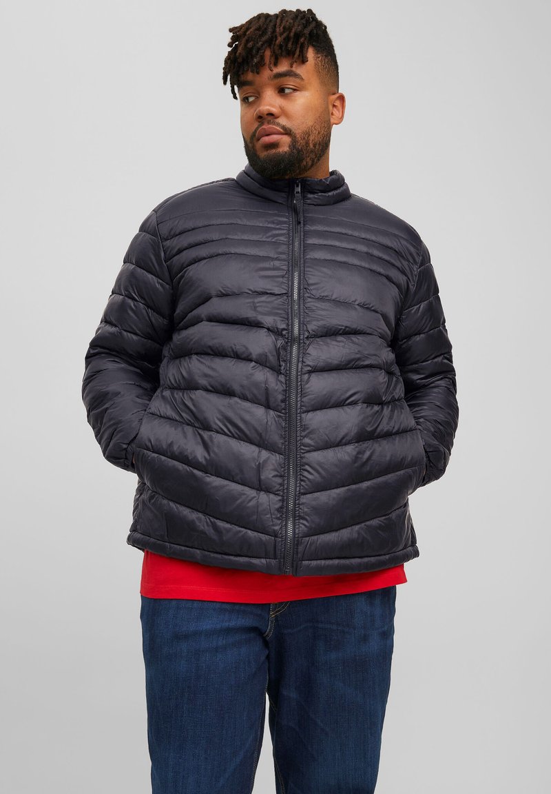 Chaqueta de plumón negra con diseño de acolchado horizontal y cuello alto. Presenta cierre de cremallera y bolsillos laterales. Llevada sobre una camisa roja.