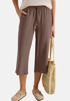 Personen har brune cropped bukser og beige slip-on sko på, og holder en vævet stråtaske ved siden.