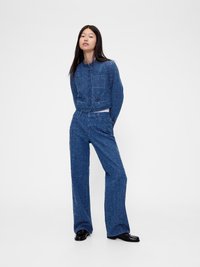 Jachetă din denim texturat albastru, cu nasturi mari și două buzunare frontale, asortată cu pantaloni din denim albastru cu croi larg. Încălțămintea este de culoare neagră.
