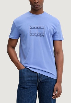 Uomo che indossa una t-shirt Tommy Jeans azzurro chiaro con logo stampato, abbinata a jeans blu scuro, in piedi con una mano in tasca.