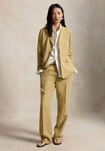 Naine seisab neutraalse taustaga, käed taskutes, kandes sinepikollast ülikonda, valget särki ja musti sandaale.