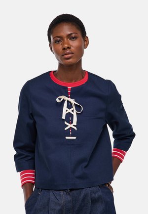 Femme portant un haut bleu marine à bordure rouge et détail à lacets blancs, associé à un pantalon bleu foncé, debout devant un fond blanc.