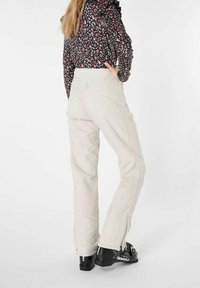 Vaaleanbeige hiihtohousut, joissa on sivuvetoketjut, istuva malli ja hienovaraiset ompeluyksityiskohdat. Yhdistetty mustiin hiihtokenkiin, joissa on kontrastivärisiä yksityiskohtia.