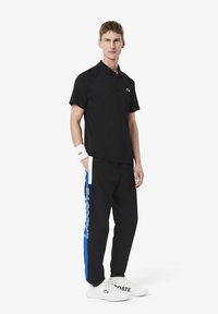 Zwarte polo met een klein groen Lacoste-logo, gecombineerd met zwarte broek met blauwe en witte "LACOSTE" tekst. Witte sneakers.