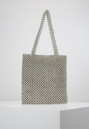 Handtas in zilverkleur met een geruit patroon van kleine metalen kraaltjes, voorzien van twee met kralen bedekte handvatten, geplaatst op een wit voetstuk.