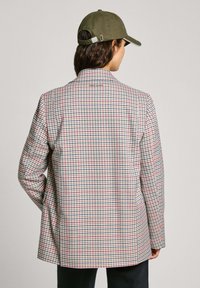 Blazer a scacchi in beige, blu e rosso. Caratterizzato da una vestibilità su misura e colletto a scialle. Indossato sopra un top scuro, con un cappello oliva.