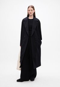 Abrigo negro largo con botones de doble botonadura, solapas anchas y corte holgado, combinado con una blusa negra y pantalones negros holgados. Bolsa tote blanca.
