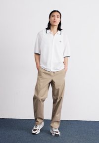 White short-sleeve shirt with a dark collar detail, beige loose-fitting trousers, and patterned sneakers standing on a blue carpeted surface.