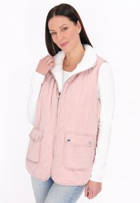 Gestepptes pinkes Westchen mit einem weißen Fleece-Kragen, zwei Fronttaschen und Reißverschluss, getragen über einem weißen Langarmshirt.