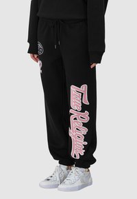 Pantalon de survêtement noir avec taille à cordon, orné d'un texte graphique rose sur le côté. Tissu en coton doux avec chevilles côtelées.