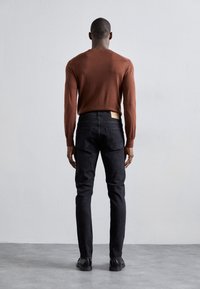 Brun stickad tröja, slim-fit svarta jeans och svarta skor. Jeansen har en läderetikett på baksidan och subtil sömnad.