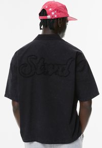 Cămașă neagră cu mâneci scurte, având un mare logo brodat "Strnd" pe spate. Prezintă un guler clasic și o croială lejeră.