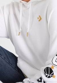 Haut blanc à capuche avec un logo en étoile dorée et une flèche sur la poitrine, graphiques noirs et dorés sur le thème du basketball sur la manche, porté avec un jean bleu.