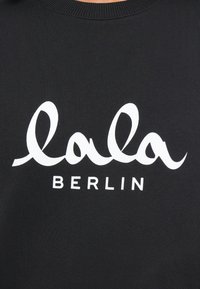 Čierna mikina s výrazným bielym textom "lala" v kurzívnom písmene a "BERLÍN" v tučnom sans-serif písme pod ním. Hladká textura.
