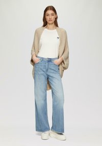 Beiger strukturierter Cardigan über einem weißen T-Shirt mit einem blauen Herzdesign, kombiniert mit hellblauen weitgeschnittenen Jeans und weißen Sneakern.