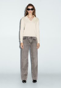 Beige pletený cardigan, šedé džíny s vysokým pasem, černé čtvercové sluneční brýle a černé boty na podpatku, s hladkou texturou a volným střihem.