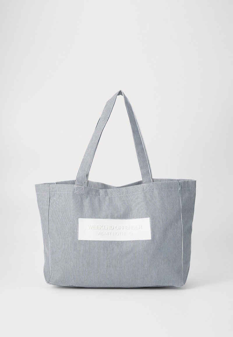 Stripete stoff totebag i marineblått og hvitt, med flat topp, to håndtak og en hvit lapp med preget tekst. Lett og romslig.
