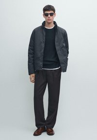 Schwarze, gesteppte Jacke über einem dunklen Pullover, kombiniert mit lockeren, dunklen Hosen und braunen Schuhen. Das Modell trägt eine schwarze Sonnenbrille.