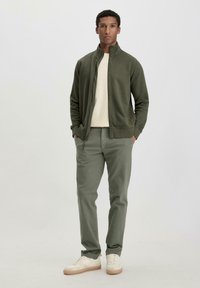 Olive green zip-up jacket over a light beige shirt, paired with light green pants and cream sneakers, standing against a neutral background.
