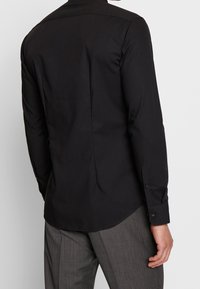 Czarna koszula z długim rękawem wykonana z gładkiej tkaniny, z prostym tyłem, mankietami zapinanymi na guziki i lekko zaokrąglonym dołem.