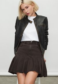 Giacca in pelle nera con collo alto, abbinata a una gonna di denim marrone scuro plissettata. La modella posa su uno sfondo neutro.