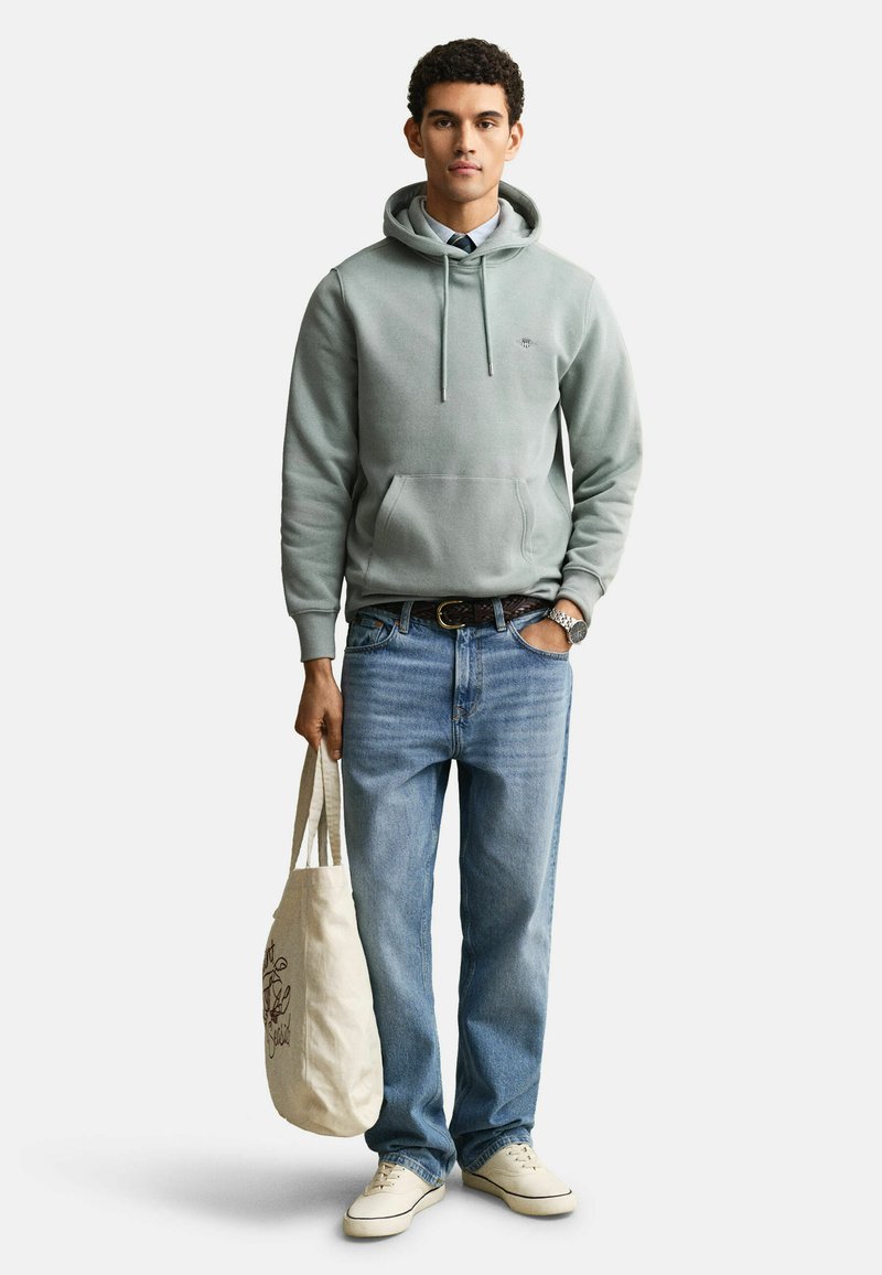 Giovane uomo con felpa con cappuccio grigio chiaro, jeans blu, sneakers bianche, che tiene una borsa tote beige con un disegno a linee nere su sfondo bianco.