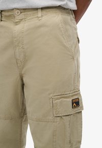 Beige cargobroek gemaakt van lichtgewicht stof, met een cargozak aan de zijkant en een gestikt logo label op de zak.