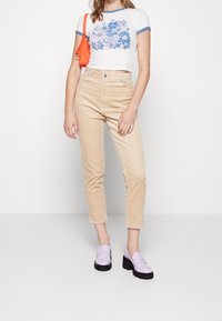 Pantalons en velours côtelé beige taille haute, t-shirt blanc court avec un imprimé floral bleu, et mocassins lavande avec des semelles noires ; sac à main orange.