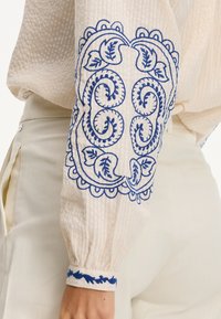 Blusa crema con tessuto testurizzato, che presenta un intricato ricamo blu sulle maniche, motivi floreali e polsini elasticizzati.