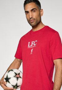 Κόκκινο βαμβακερό T-shirt με εκτύπωση "LFC" και "ANFIELD RD, LIVERPOOL, L4" σε λευκό χρώμα. Το μοντέλο κρατά μια ασπρόμαυρη μπάλα ποδοσφαίρου με σχέδιο.