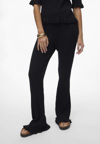 Pantalons noirs à côtes avec une jambe évasée et un ourlet froncé. Associés à des sandales à plateforme marron et à un bracelet en or au poignet.