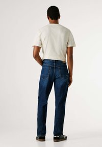 Vaqueros de mezclilla azul con corte recto y dos bolsillos traseros. El modelo lleva una camiseta de color beige claro y zapatillas negras.