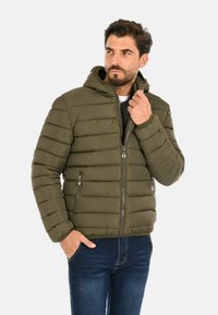 Uomo che indossa una giacca a cappuccio trapuntata verde oliva e jeans blu, mentre aggiusta la cerniera della giacca con una mano e tiene l'altra mano in tasca.