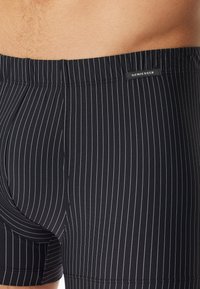 Ingrandimento di una persona che indossa un costume da bagno nero con sottili righe verticali bianche e una piccola etichetta nera "Schiesser" sulla vita.