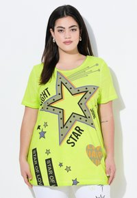 Lysende grøn T-shirt med et stort stjernegrafik, omkranset af grå og orange, med tekst og mindre stjernemønstre. Korte ærmer, afslappet pasform.