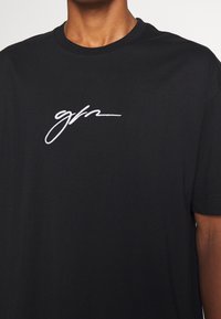 Svart bomull t-shirt med en vit broderad signaturlogotyp på bröstet, korta ärmar och rund halsringning.