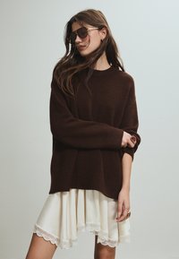 Maglione marrone, oversize, con maniche lunghe e struttura morbida. Abbinato a un vestito bianco con bordo in pizzo all'orlo. Occhiali da sole scuri indossati.