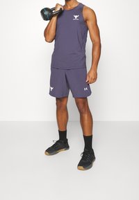 Man i lila ärmlös tröja och shorts håller en kettlebell på axeln, iförd svarta strumpor och svarta träningsskor.