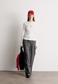 Cămașă albă cu mâneci lungi și text, pantaloni largi gri cu dungi, șapcă roșie și pantofi negri. Modelul ține o jachetă roșie și o geantă neagră.